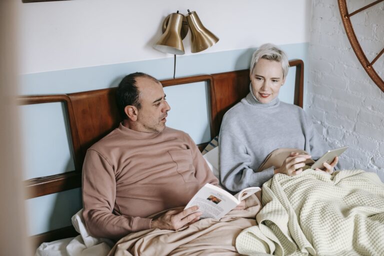 Middle aged ethnic man in casual clothes sitting on bed and reading book near positive wife using tablet