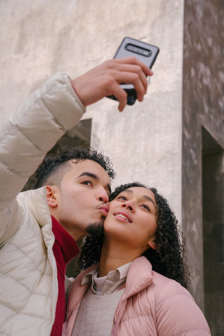 Romantic ethnic man kissing cheek of girlfriend and taking selfie