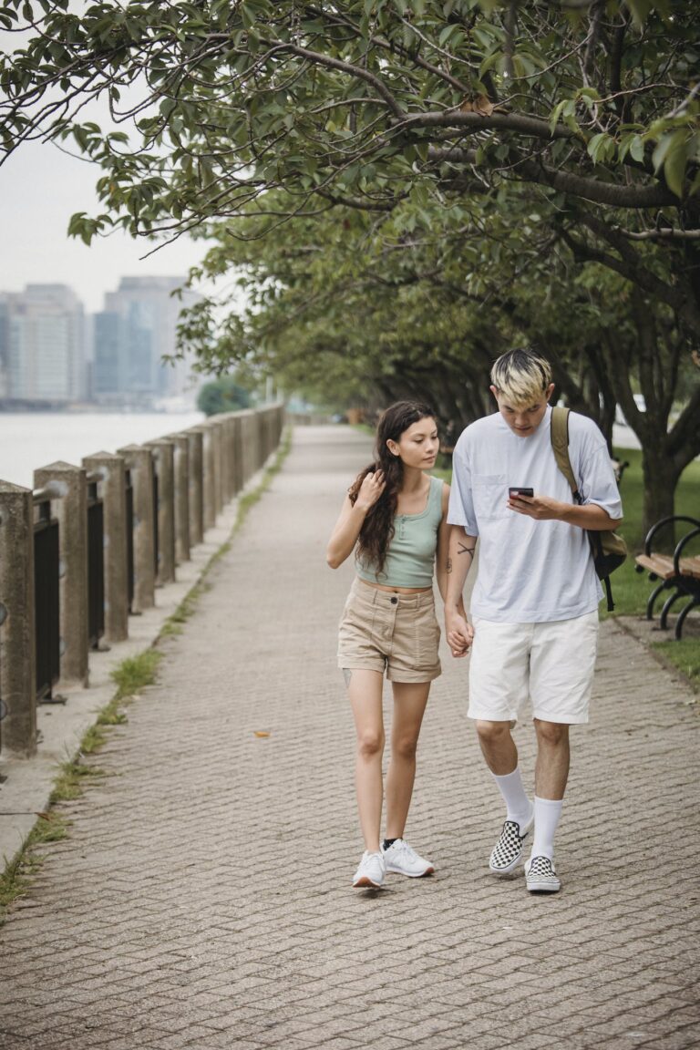 Young stylish couple using smartphone while strolling in park at riverside